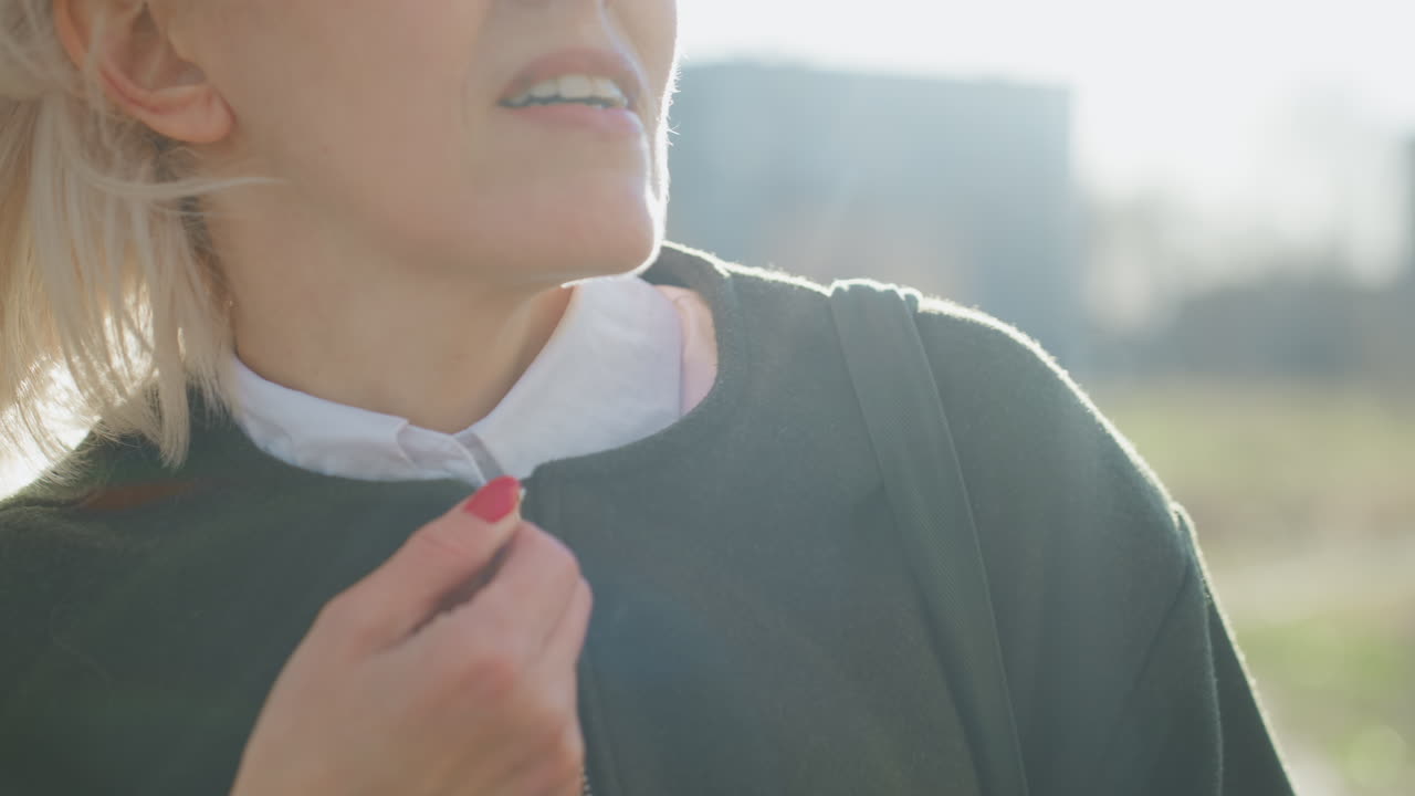 Stylish adult woman outdoors wearing sunglasses and dark jacket, blonde hair blowing across face in strong wind, white shirt visible, candid natural moment with blurred background of grass and trees