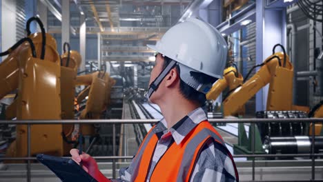 close up side view of asian male engineer with safety helmet taking note on the tablet and looking around while standing with industrial robotic arms working