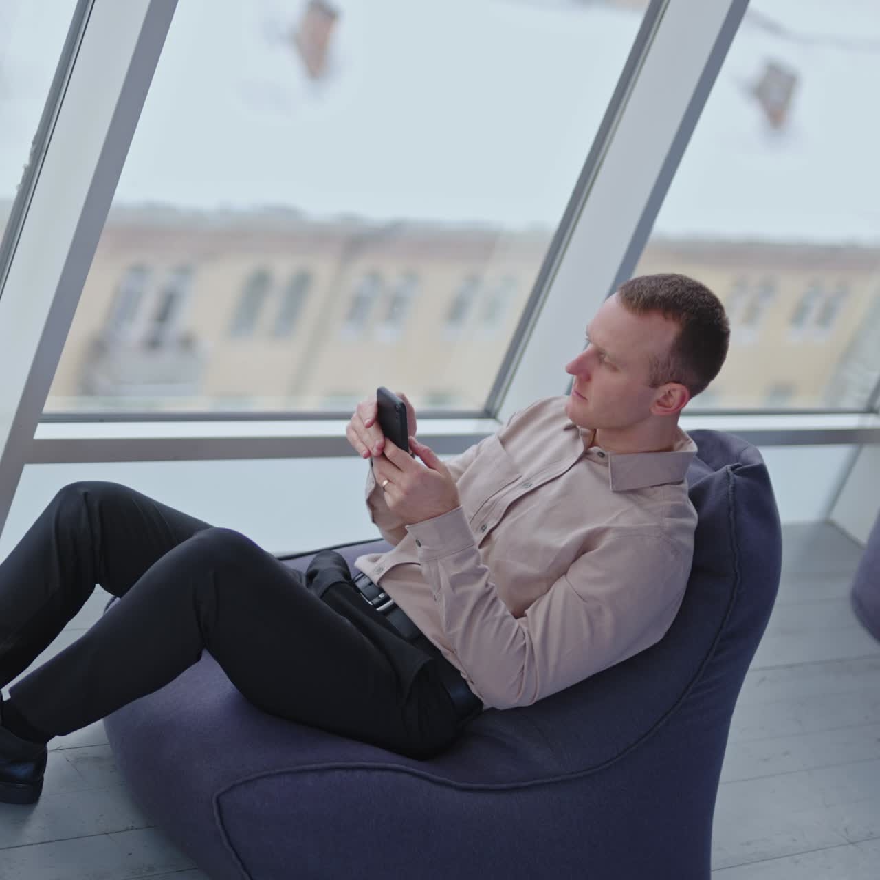 Male worker resting in the bean bag chair. Man searches information on the phone. Panoramic window with city view at backdrop