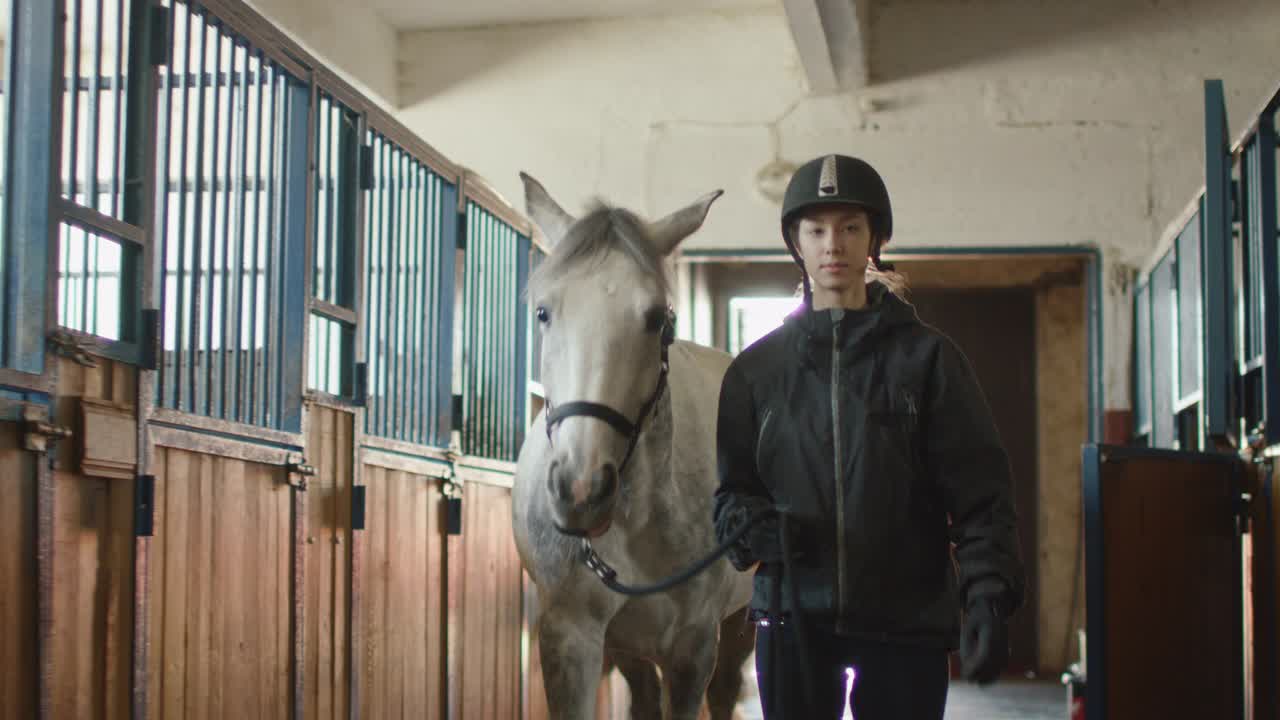 una joven jockey está caminando con un caballo blanco en un establo.