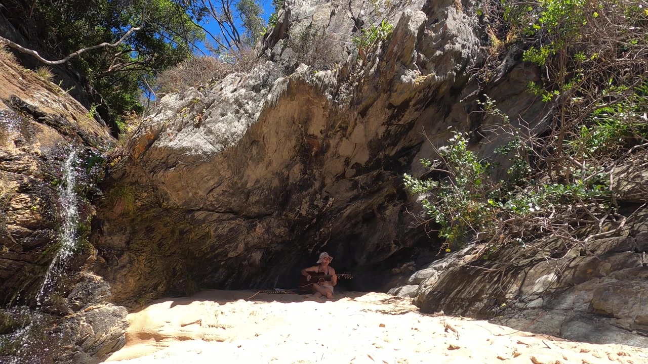 En ensom kvinde sidder på en sandstrand under en udhængende klippe og spiller guitar nær et blidt vandfald, der fosser ned ad klippeklipper, ved Storm’s River Mouth, Tsitsikamma Nationalpark, Sydafrika.