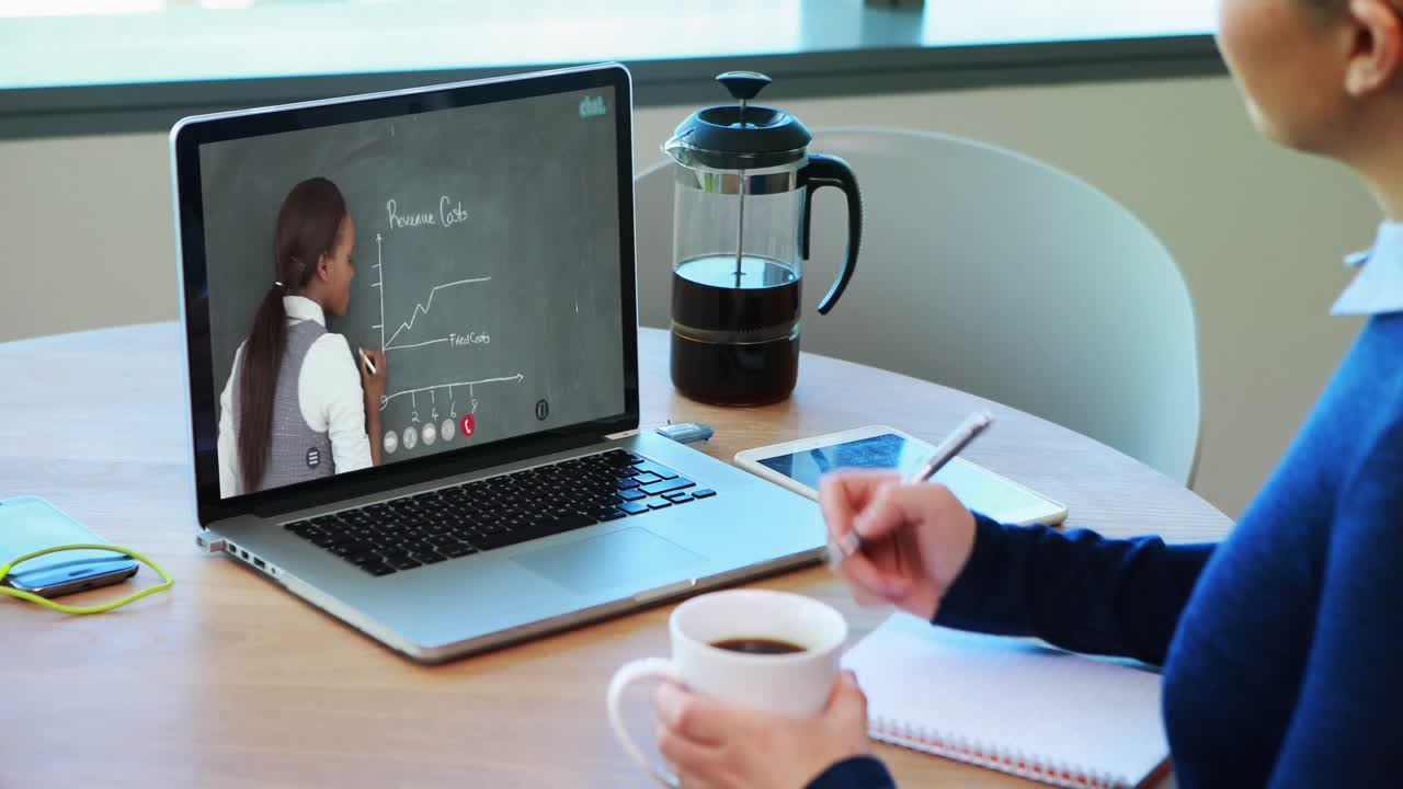 Caucasian female student using laptop on video call with female teacher, making notes