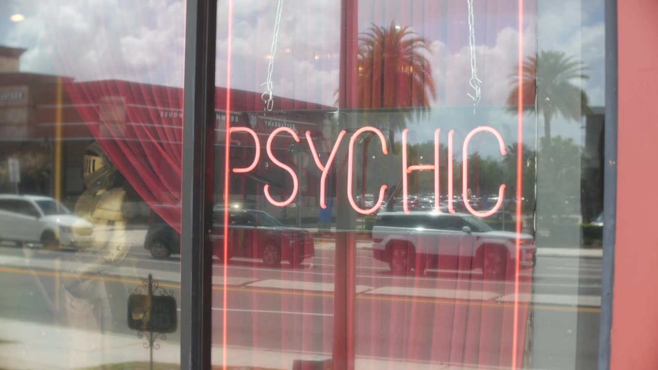 Psychic neon sign glowing in a storefront window with street reflections. Cars, palm trees, red curtains, and bright daylight create an urban retail scene.