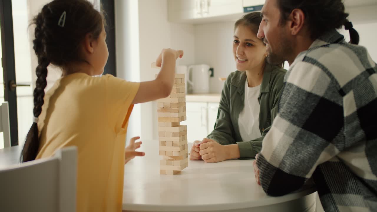 Rear view of a little brunette girl in a yellow dress playing Jenga with her parents in a white kitchen in a modern apartment. A little girl plays a game for developing hand motor skills with her parents and communicates with them while sitting in the kitchen at home