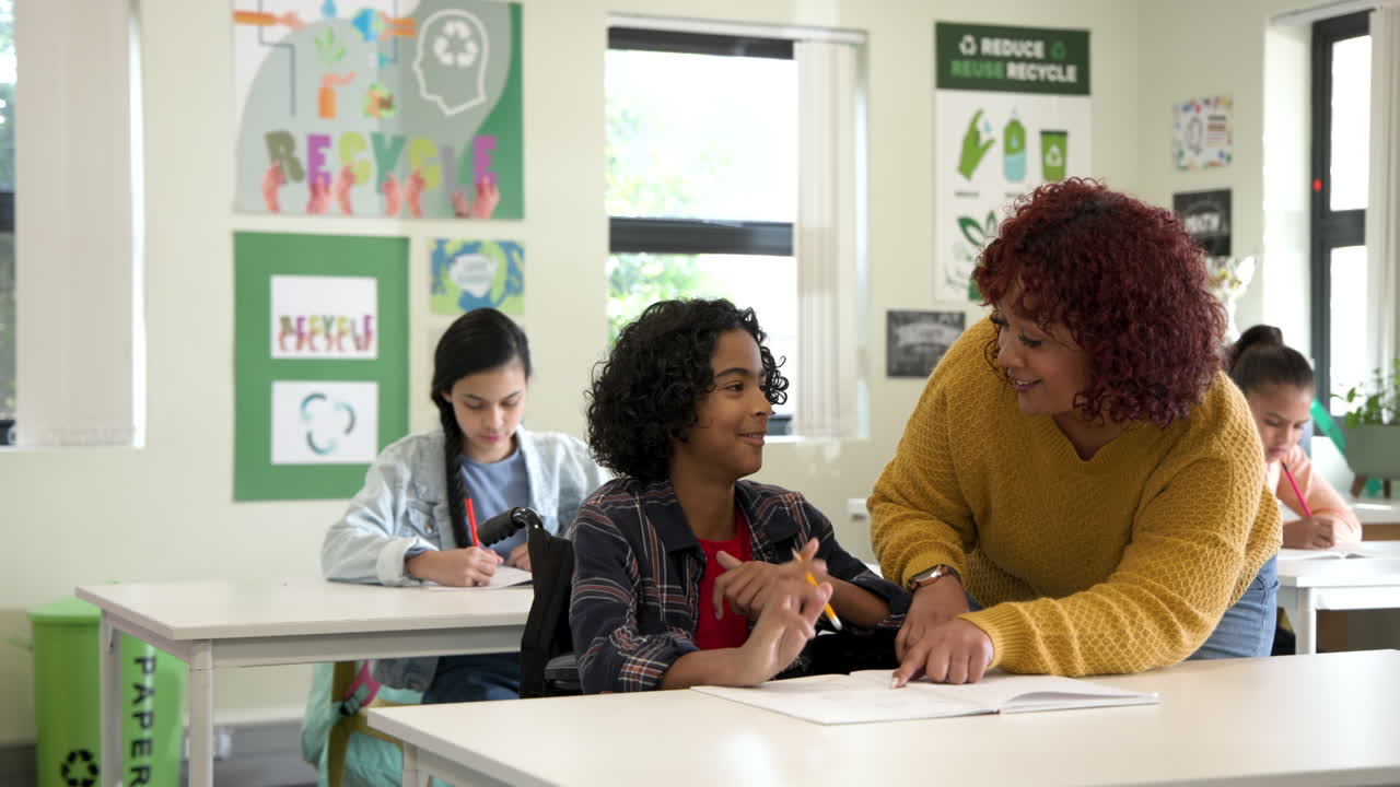 Female teacher helping boy in wheelchair with writing in classroom, fostering learning, at school