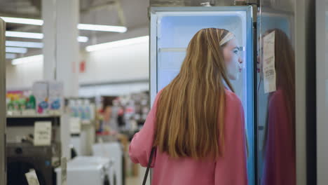 lady opens glowing refrigerator in a brightly lit store, carefully inspecting the illuminated interior, the refrigerator's light enhances the sleek, modern design as she holds the door