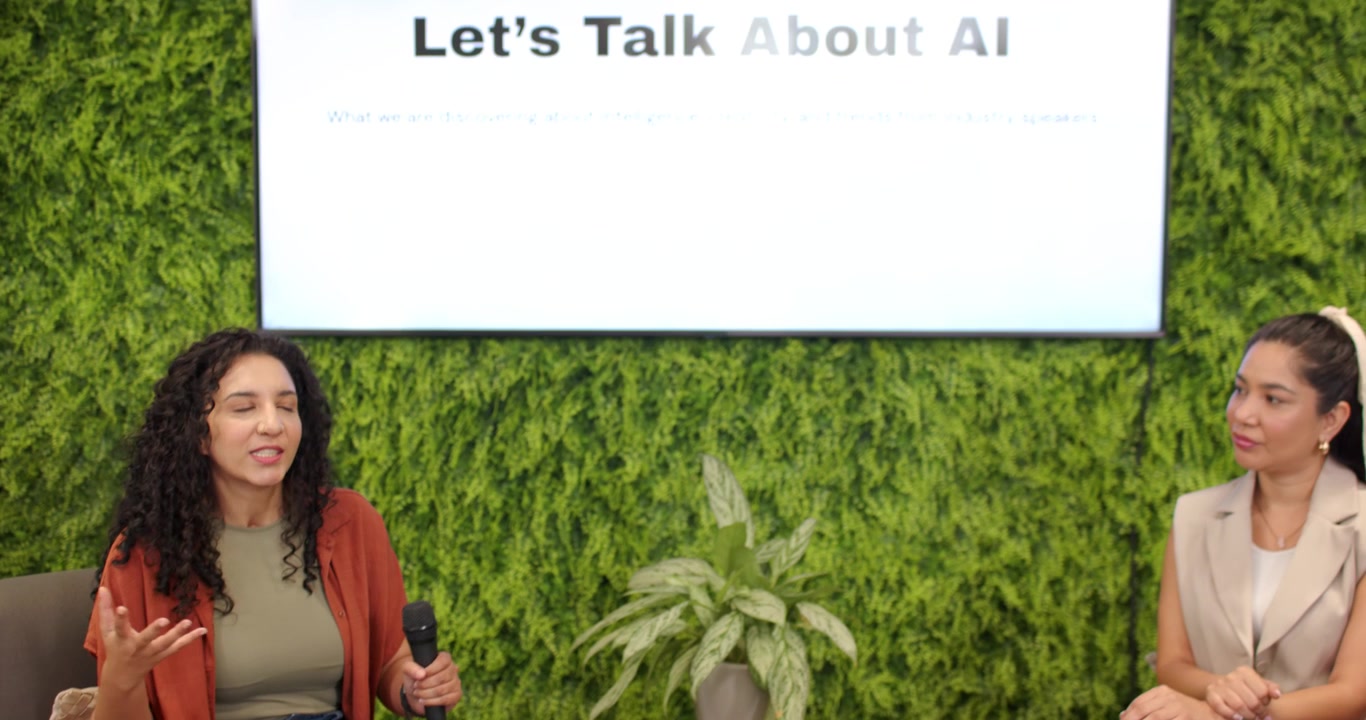 Female panelists holding microphones and discussing AI in office lounge being prompted by slide