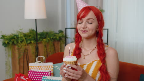 happy redhead girl celebrating birthday party, makes wish blowing burning candle on small cupcake