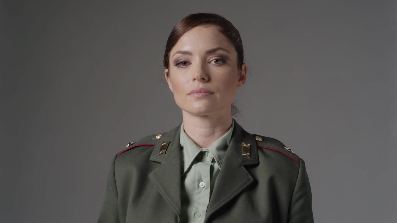 Standing officer in green jacket making subtle face shifts during studio zoom, showing authority