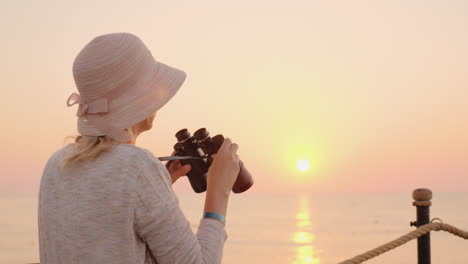 Una-Joven-Mujer-Mirando-A-Través-De-Binoculares-En-La-Distancia-Para-Cumplir-Un-Sueño-Rosa-Se-Sienta-En-Un-Muelle-De-Mar-En