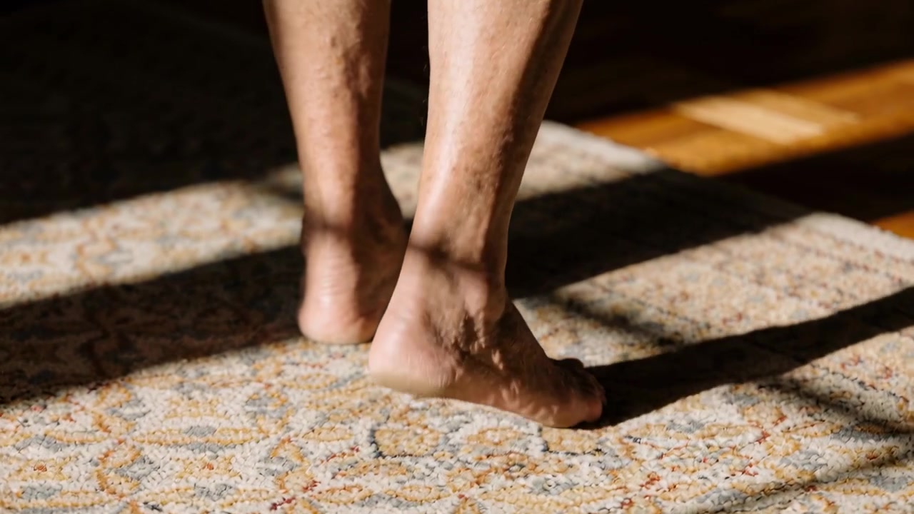 Gambe e piedi nudi che si muovono verso il parquet caldo su un tappeto a motivi, sotto le ombre della finestra, in cerca del sole.