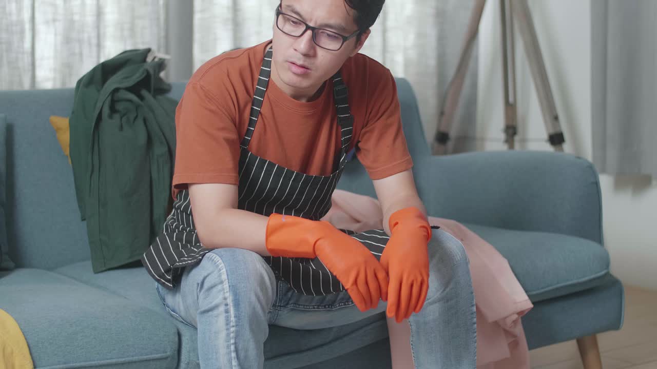 ama de llaves masculina asiática con un delantal sentada en un sofá y con dolor de cabeza debido a estar cansada limpiando la habitación desordenada