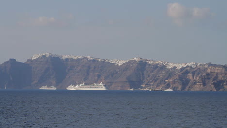 una vista de los majestuosos acantilados negros de santorini con un pueblo construido al borde de ellos, cruceros estacionados justo debajo