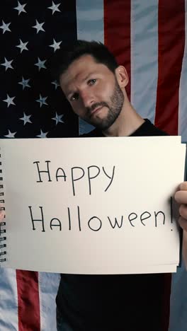 happy and confident tattooed man lifting up a notepad that says happy halloween on it in black writing on white paper. man is standing in front of an vertical american flag that is slowly moving
