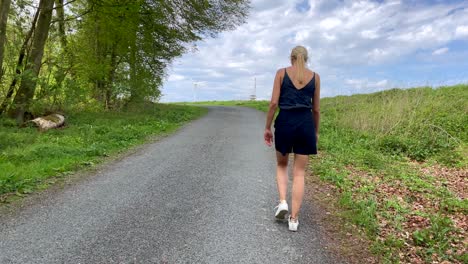 slow motion shot of pretty girl in shorts walking on asphalt path surrounded by green nature with forest and trees during sunny and cloudy summer day