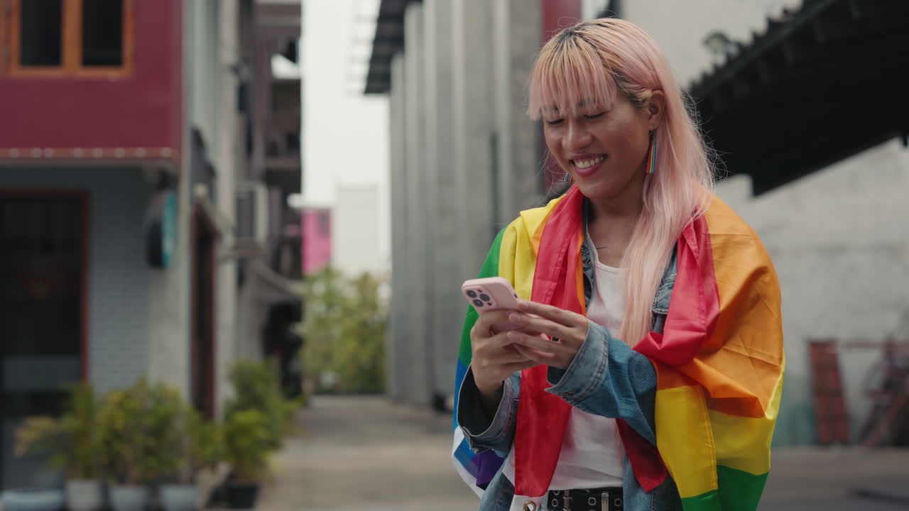 Woman using mobile phone, draped in pride flag, in urban setting