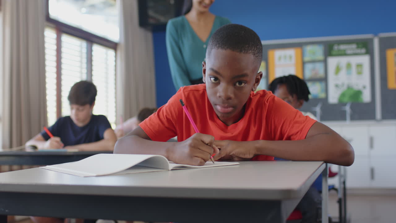 Smiling boy in school classroom writing in notebook with classmates and teacher