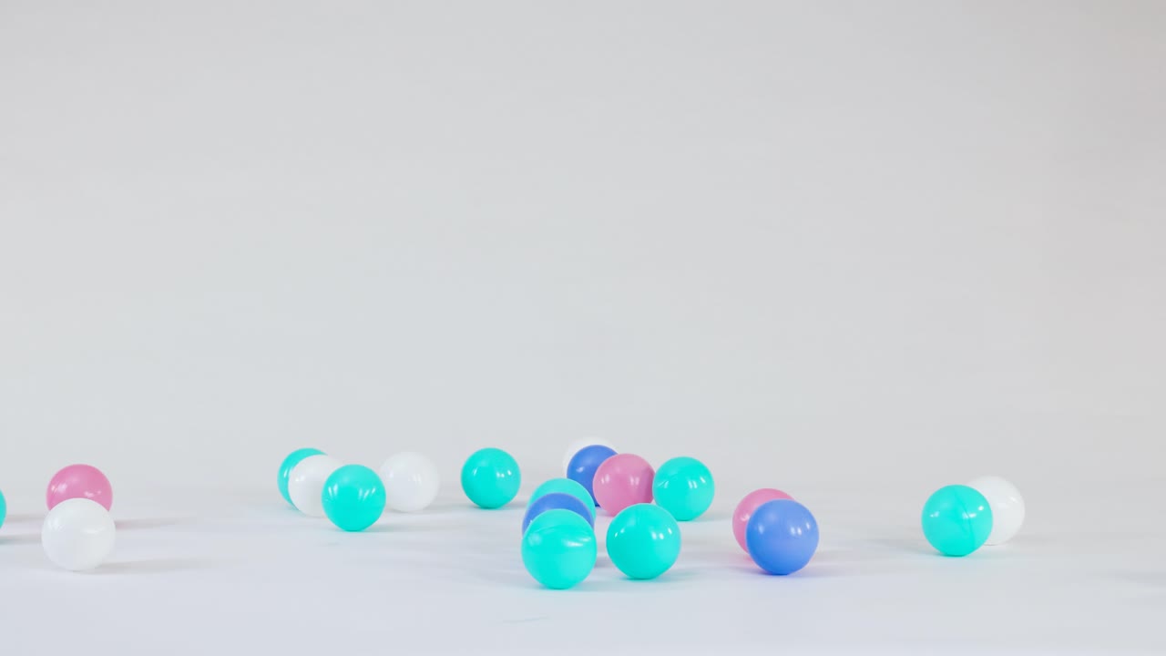 Pink, blue, and white plastic spheres fall and bounce across a clean white studio floor