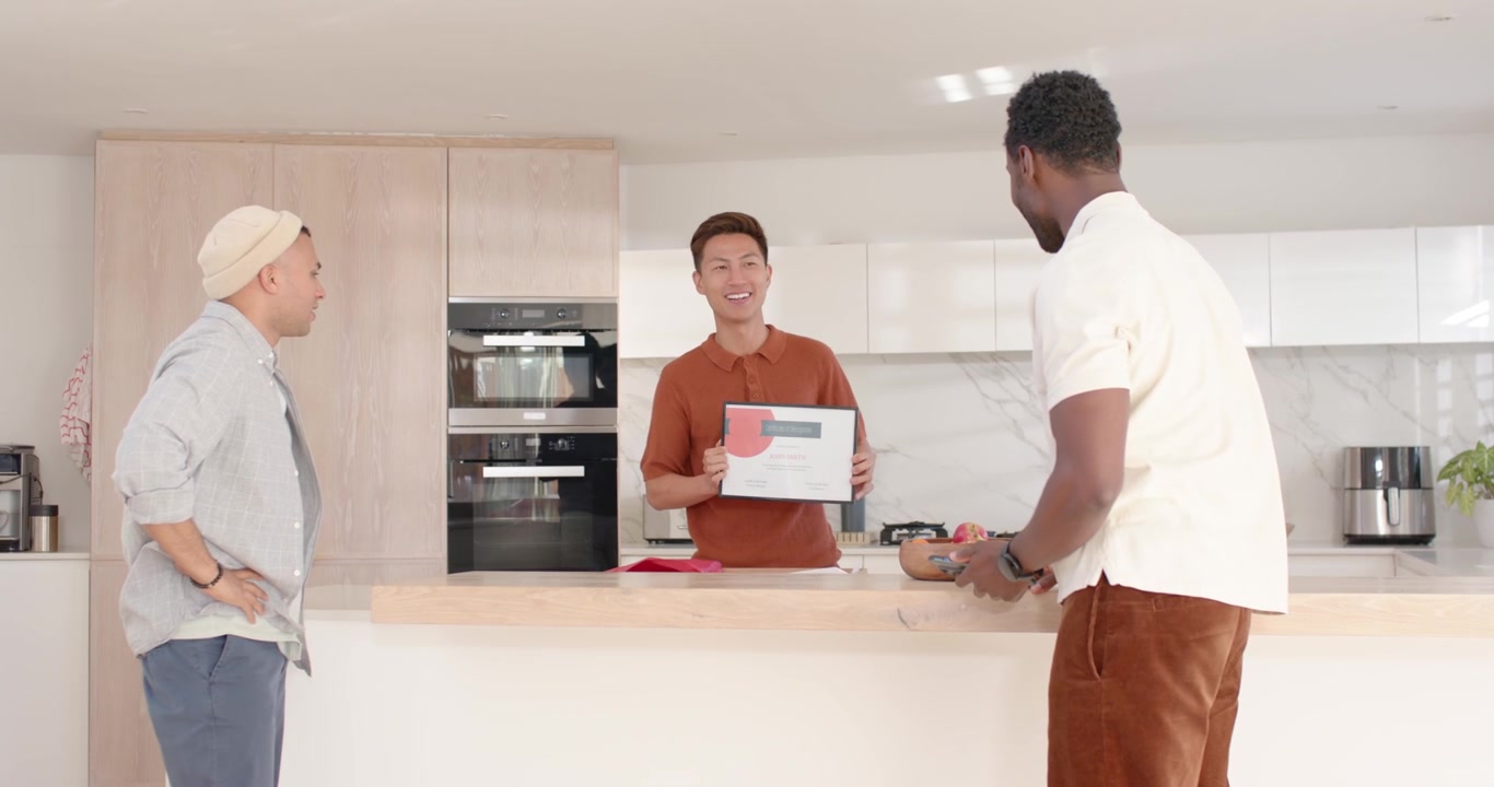 Diverse male friends presenting framed certificate, shaking hands and celebrating at kitchen island