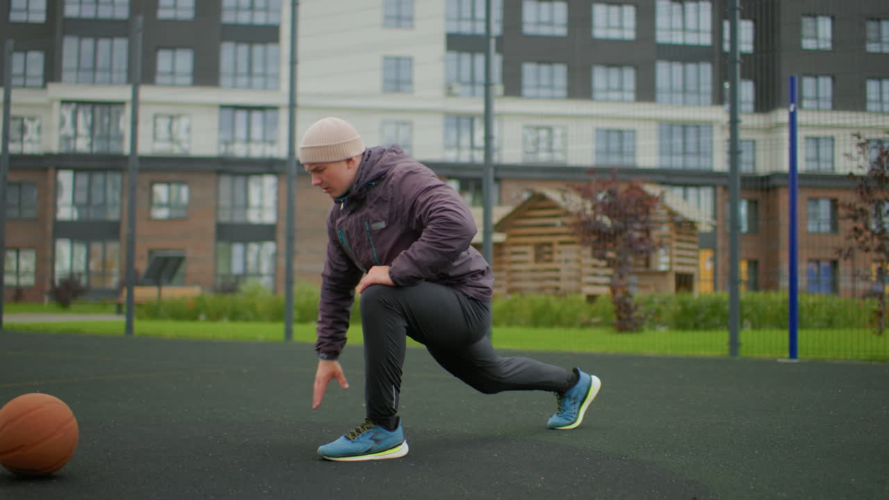 Senior caucasian man stretching on court, performing deep lunges and dynamic reach with basketball nearby, wearing beanie and jacket, cloudy morning, apartment blocks in background, former coach
