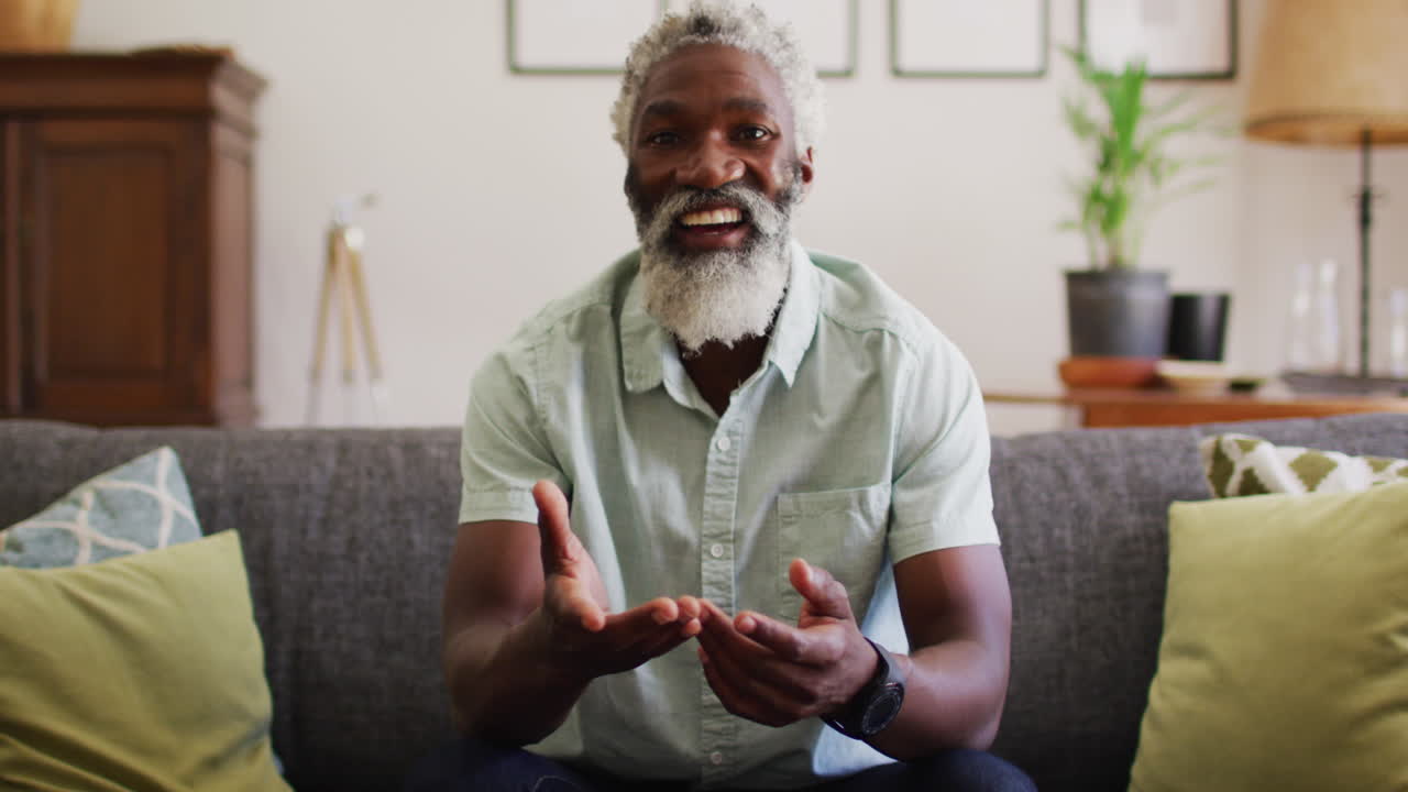 Happy african american senior man sitting on couch making video call smiling and waving