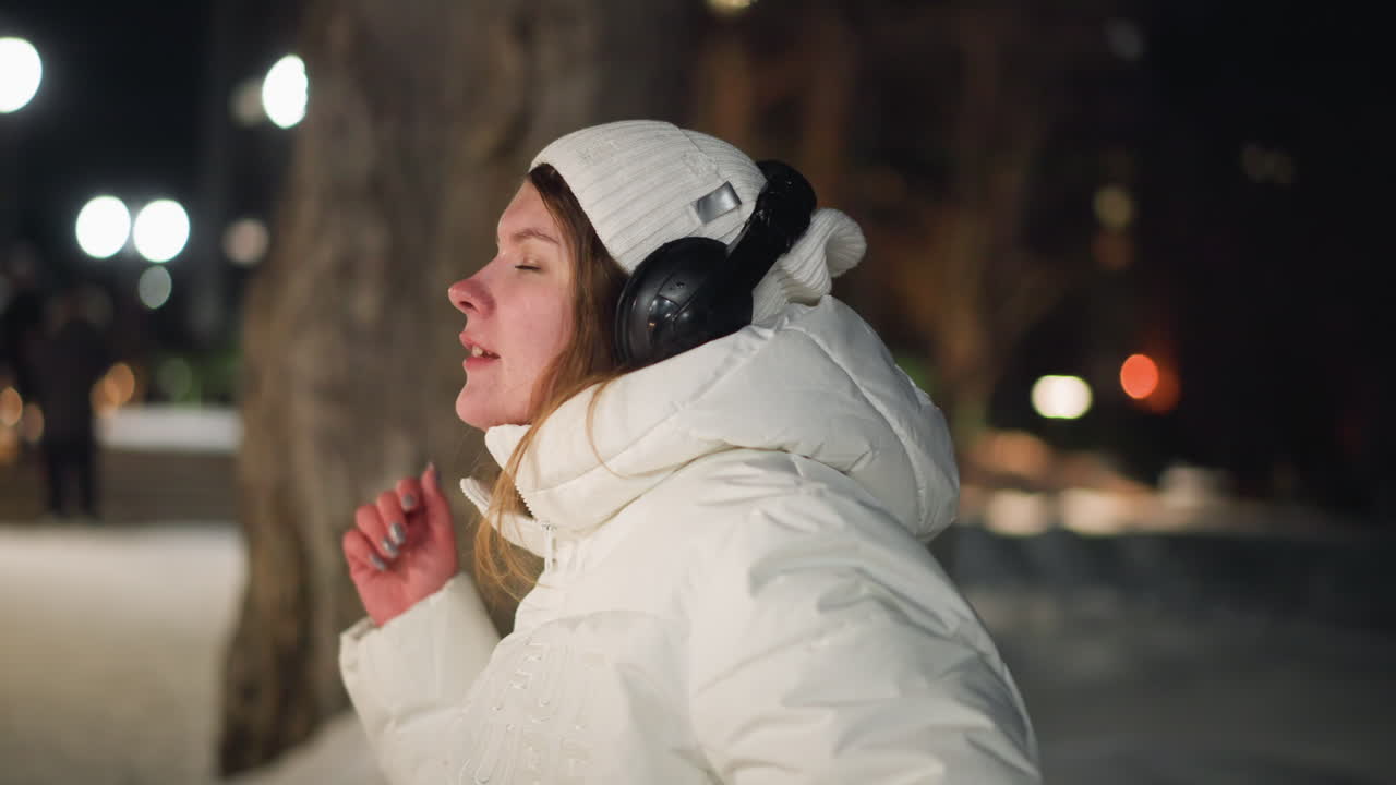 Female artist seated in snowy park at night, wearing white winter coat and headphones, eyes closed in peaceful moment, enjoying music while people pass by in well lighted outdoor environment
