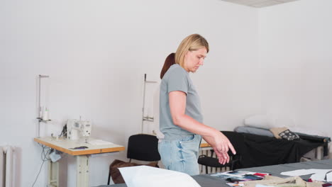 weavers walking towards table in creative workspace while engaging in conversation, one trying to match her textile with cloth, sewing machine and workspace equipment visible in background