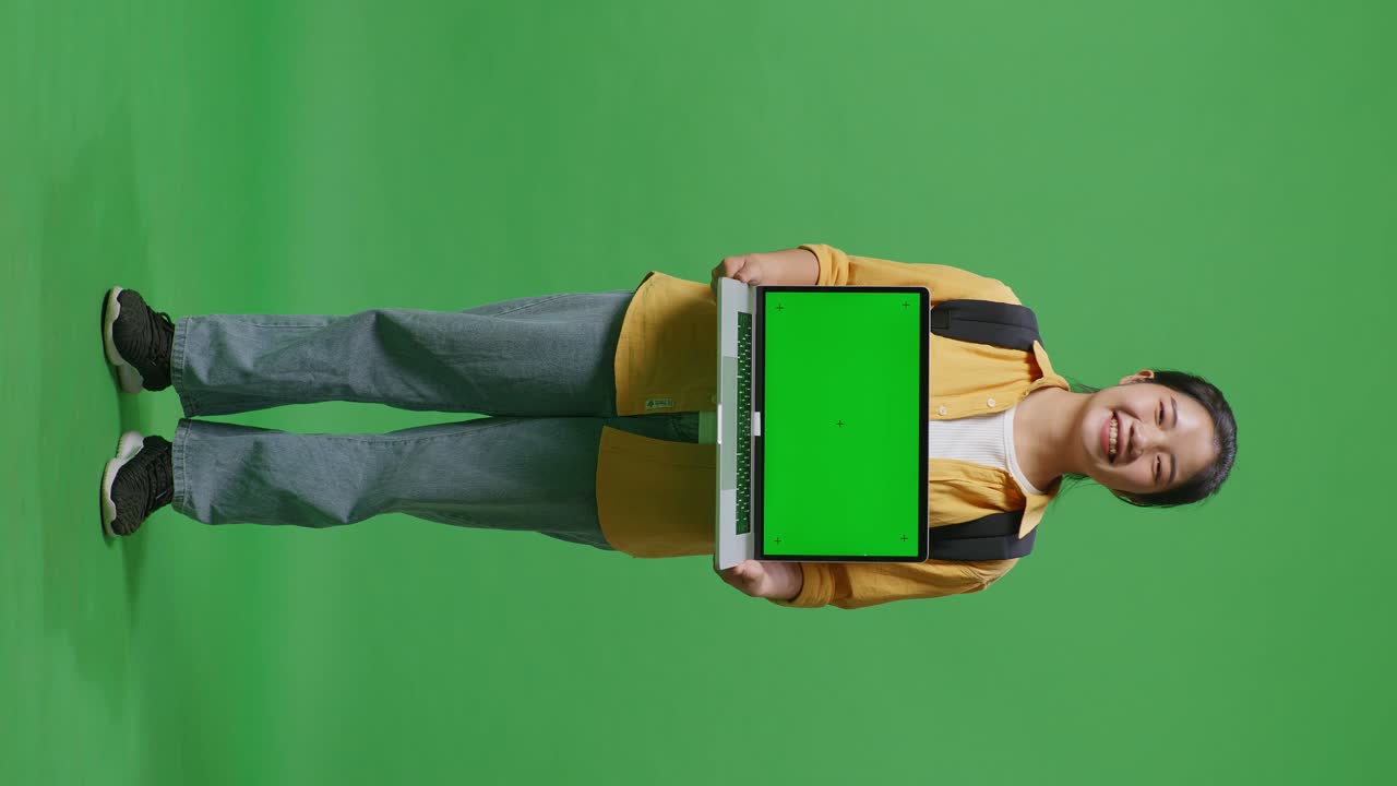 cuerpo lleno de estudiante mujer asiática con una mochila sonriendo y mostrando pantalla verde portátil a la cámara mientras estaba de pie en la pantalla verde estudio de fondo