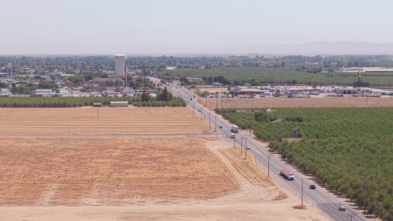 Aerial reverse shot following road between fields, leading toward city and water tower in Ripon California