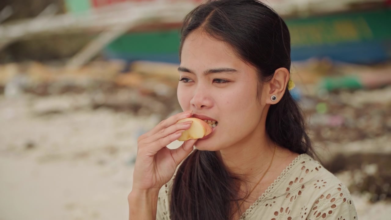 Closeup asian woman eating lunch on beach, focused facial expressions and long dark hair, embroidered blouse, holding foam container and taking bite, pausing to reflect, driftwood and small boat