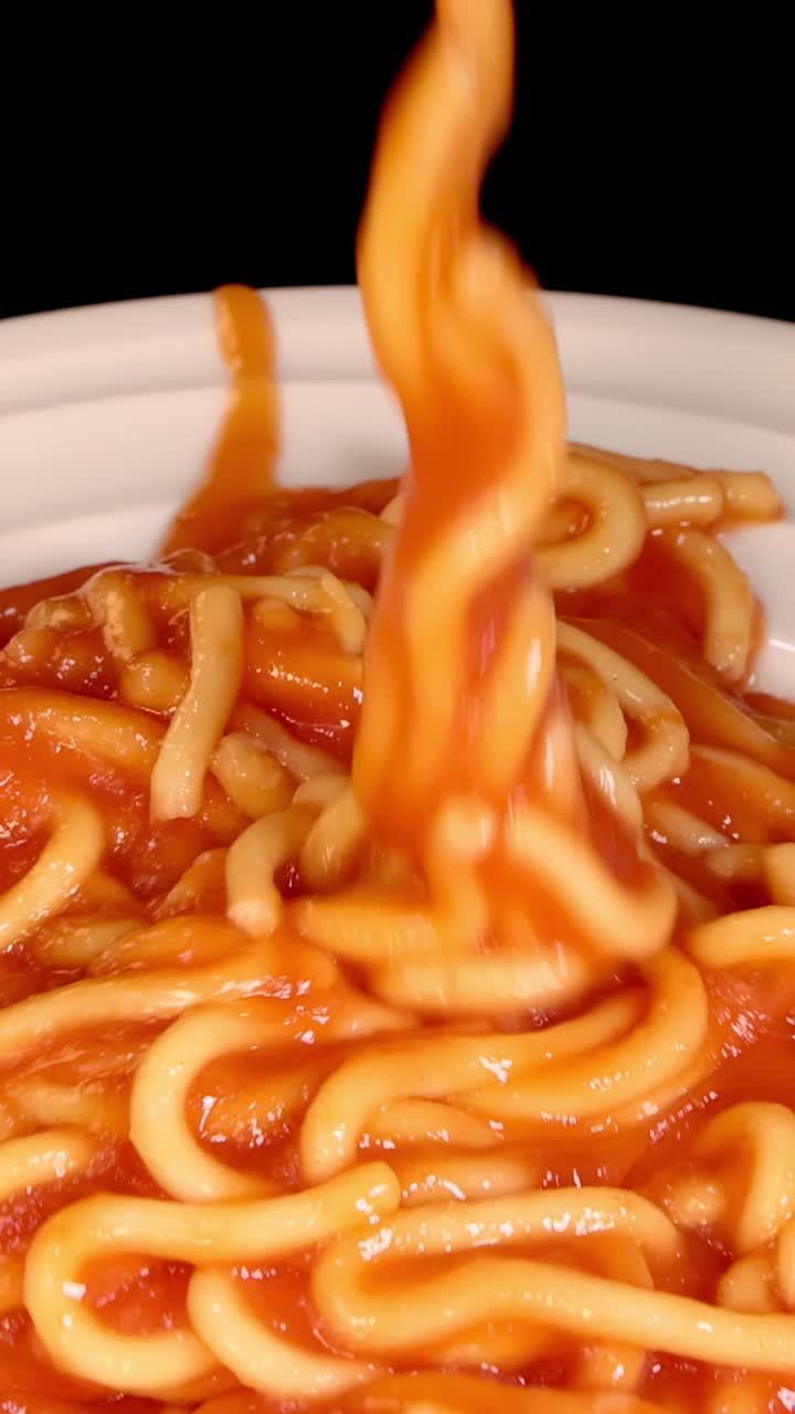 Spaghetti cotti con salsa di pomodoro vengono fatti cadere su un piatto bianco pulito, ripresi in primo piano con illuminazione brillante e uniforme e una telecamera statica.