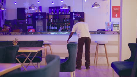 a person cleaning up bar counter in stylish modern lounge with bottles on shelves, purple lighting, and contemporary seating in background. relaxed indoor setting for hospitality services