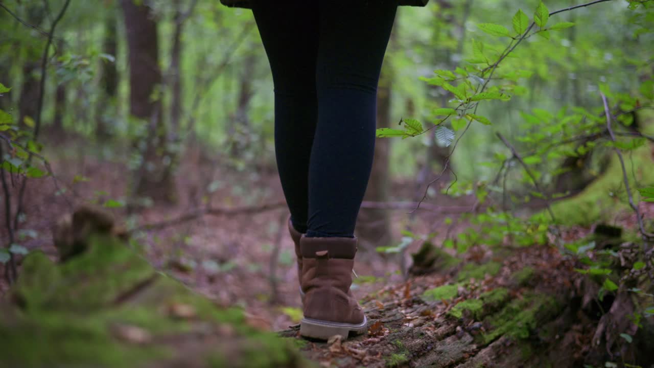 Primo piano di una persona che indossa scarponi da trekking marroni mentre cammina su un albero caduto coperto di muschio in una lussureggiante foresta verde. Il video evoca un senso di esplorazione, connessione con la natura e avventura all'aria aperta.