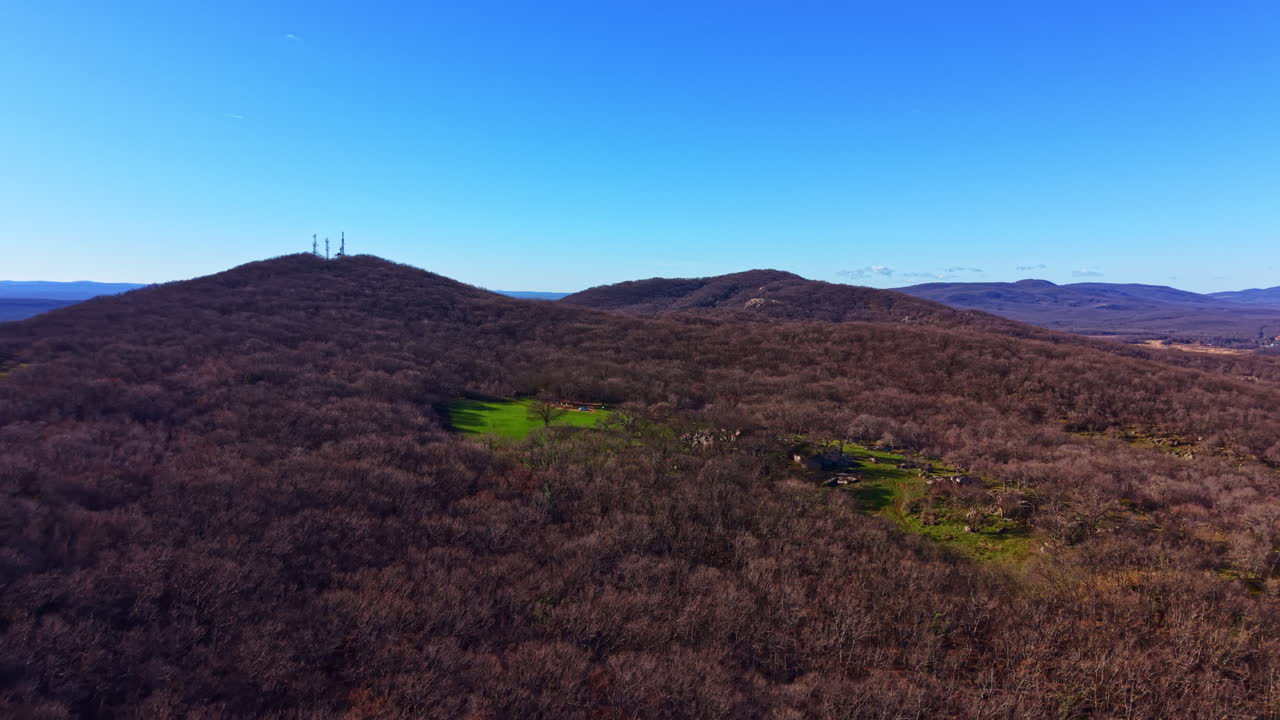 Udsigt over en skovklædt bjergside med kommunikationstårne på toppen under en klar himmel i dagslys