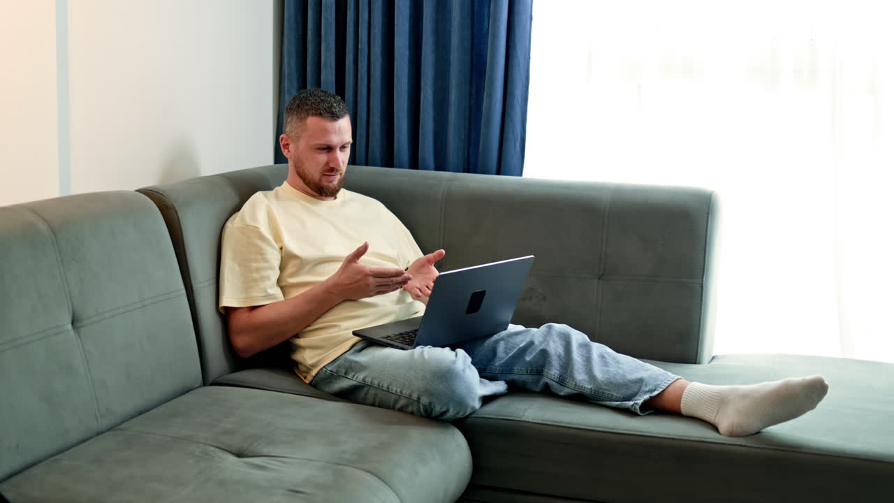 Man sits on couch using laptop for a video call while speaking and expressing gestures with hands in living room during daytime
