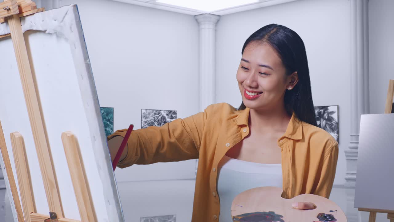 아시아 여성 예술가 가 갤러리에서 오일 페인트와 브러쉬로 캔버스에 그림을 그리는 동안 카메라에 미소 짓고 있습니다.