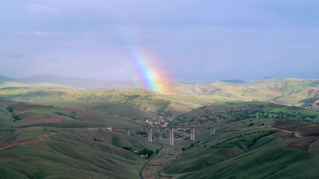 Duha nad údolím s mostem a malou vesnicí.
