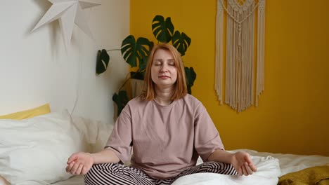 woman sitting on bed meditating with closed eyes in lotus pose