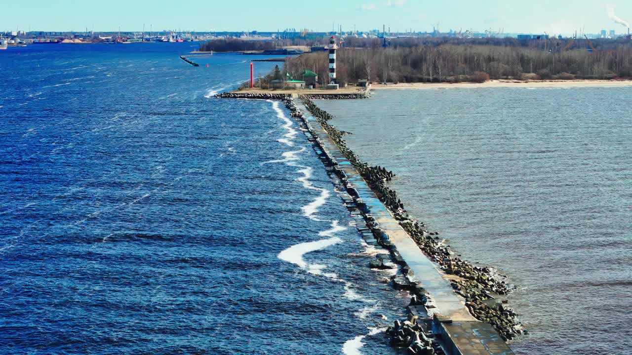A long stone jetty stretches into bright blue Baltic Sea, ending at a black and white lighthouse that marks the split from tranquil estuary waters and sandy forested shore.