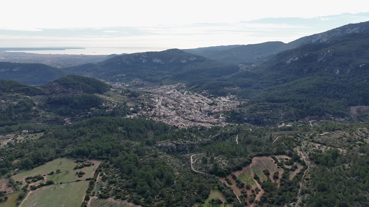 Volo aereo all'indietro sopra le verdi colline di Maiorca e il villaggio di Esporles nella valle durante il giorno.