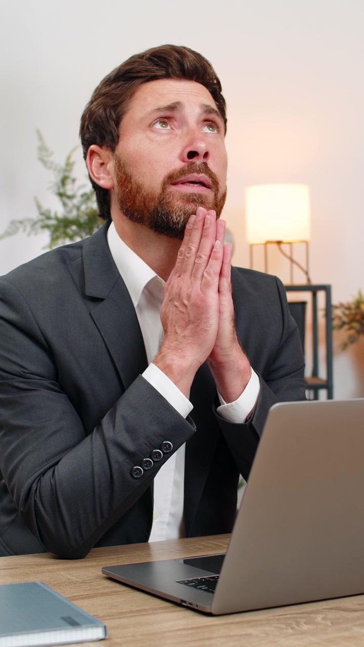Uomo d'affari di mezza età giunge le mani in preghiera davanti al laptop, guarda in alto desiderando un ordine dal cliente o l'approvazione del direttore.