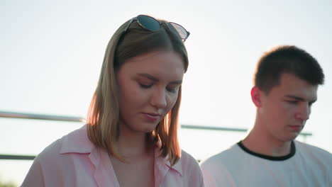 close-up of young woman with glasses resting on head illuminated by warm sunlight creating a glowing halo, standing beside a focused young man outdoors with railing