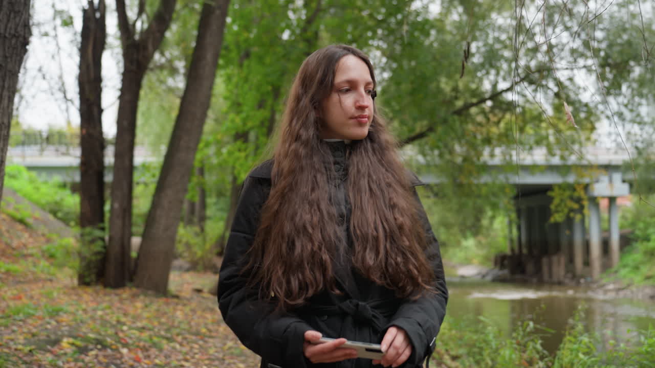 caucasian woman walking in park, long brown hair and black coat, holding phone near river bank under tall trees, bridge and urban background, autumn leaves on ground, contemplative expression, slow