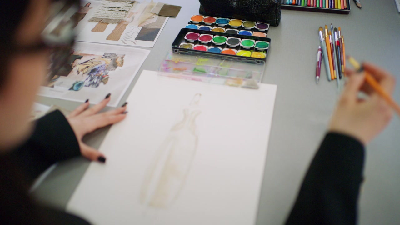 Close up of a hand painting a fashion figure sketch while a watercolor palette, paint tray, brushes, pen, and glass of water sit nearby