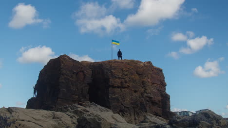 Menschen-Klettern-Und-Stehen-An-Einem-Sonnigen-Tag-Auf-Dem-Kapellenfelsen-Mit-Ukrainischer-Flagge,-Perranporth,-Cornwall,-England,-Großbritannien---Zeitraffer