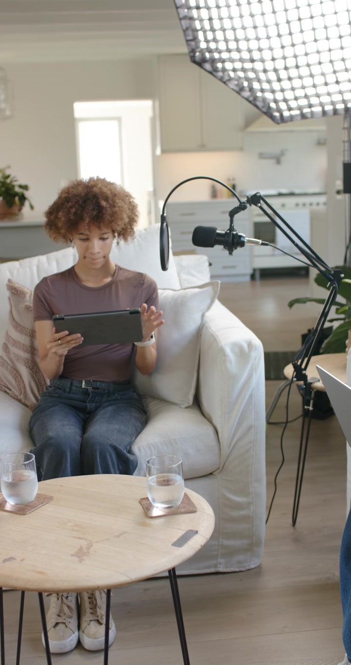 Vertical video: Women typing at laptop on sofa tapping tablet camera panning prepping podcast mic