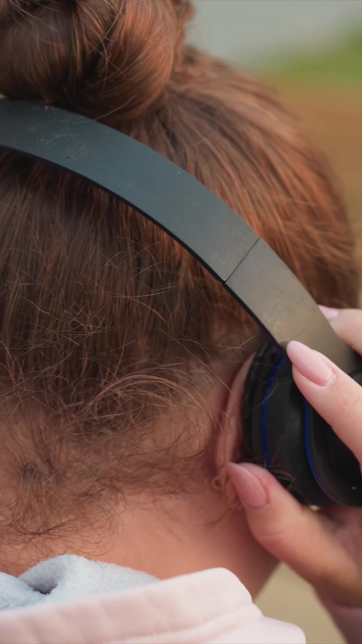 Soggetto visibile da dietro mentre rimane fermo. Una mano sistema le cuffie nere over-ear sul lato destro della testa. Vegetazione visibile ma non identificabile oltre la forma generale.