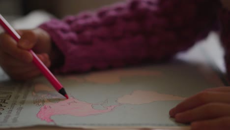 a toddler girl is coloring the continent of north america with a pink pencil in a world atlas educational book