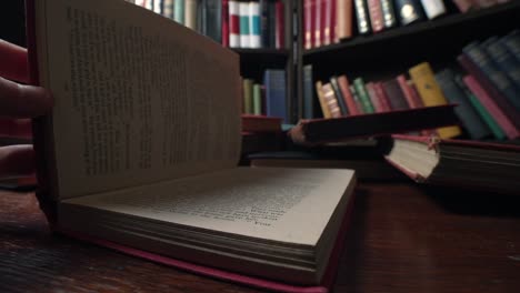 Hand-shuts-book-with-red-cover-in-library,-many-books-on-table-behind,-macro-view