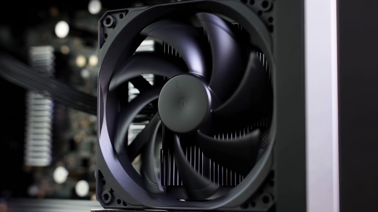 A close-up view of a black cooling fan spinning inside a desktop computer case, with internal hardware softly blurred in the background. Centered framing, low-key lighting, and visible motion create a technical and modern atmosphere